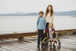 Three people stand on a wet wooden boardwalk beside a calm body of water with misty hills in the background. An adult stands behind two children, with one child standing and another seated in a yellow mobility walker. A wooden railing runs along the boardwalk, and the overcast sky creates soft, diffused lighting throughout the scene.