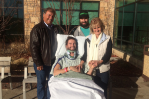 A smiling young man, Mike Shaw, sits in a supportive medical chair outdoors wearing a hospital gown and a cervical neck collar. He is surrounded by three supportive family members—an older man in a leather jacket on the left, a man wearing a black baseball cap in the back, and a woman in a white vest holding Mike's hand on the right. They are all smiling together outside a brick and glass hospital building on a sunny day.